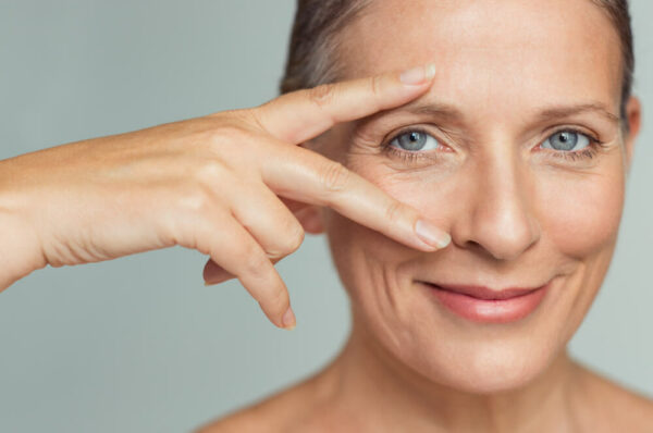 Portrait of smiling senior woman with perfect skin showing victory sign near eye on grey background. Closeup face of mature woman showing successful results after anti-aging wrinkle treatment.